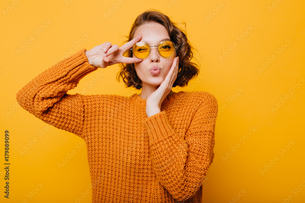 Stunning short-haired female model posing with kiss face expression on yellow background. Close-up portrait of stylish european girl standing with peace sign in front of wall. - obrazy, fototapety, plakaty 