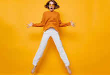 Full-length Portrait Of Funny Woman In White Pants Fooling Around On Yellow Background. Barefooted Curly Girl In Sunglasses And Soft Sweater Jumping And Waving Hands With Shocked Face Expression.
