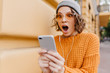 Shocked girl with brown eyes looking at phone screen with mouth open. Outdoor portrait of surprised young woman with curly hair wearing yellow knitted attire and holding smartphone.