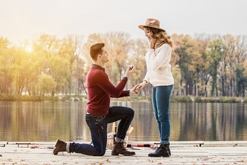 Marry me. Horizontal shot of young man in casual clothing standing on one knee and holding engagement ring while making wedding proposal to his beautiful girlfriend. Engagement of young couple.