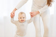 © Rithor - Cute little baby is looking at camera and smiling while learning to walk, mom is holding his hands. first steps of child. White light domestic interior, white clothes