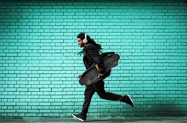 Handsome young dreadlocks skater with headphones running in a suit near the blue wall on the street.