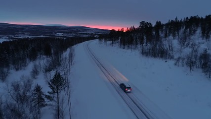 Wall Mural - AERIAL: Flying above car driving along empty icy mountain highway through snowy spruce forest at golden winter sunset. People on road trip traveling across snow covered Lapland wilderness at sunrise