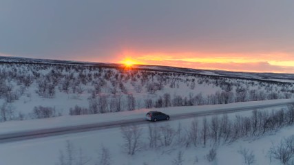 Wall Mural - AERIAL Flying above blue car driving along slippery snowy highway past frosty bare trees at golden sunrise. People on road trip traveling across snow covered Lapland wilderness at orange winter sunset