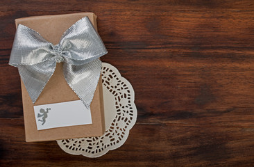 Wall Mural - Wrapped gift box with silver ribbon bow on a dark walnut wood, on lace napkin with white card and silhouette of angel, top view, close up