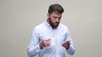 Poster - Surprised bearded man in shirt using smartphone while drinking tea over gray background