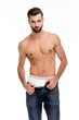 © MARIIA - Time to undress. Handsome shirtless young man looking at camera and taking off his jeans while standing against white background.