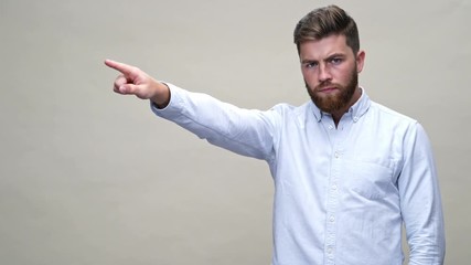 Canvas Print - Serious bearded man in shirt with crossed arms pointing at someone and expels him over gray background