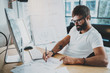 © SFIO CRACHO - Bearded coworker professional wearing eye glasses working at modern loft studio-office with desktop computer.White blank display screen.Blurred background. Horizontal.