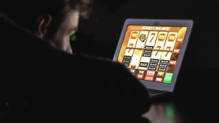 Gambling addicted man in front of online casino slot machine on laptop computer at night - loosing his money. Dramatic low light grain shot.