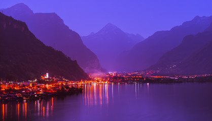 Wall Mural - Lights of the night city. Switzerland. Night panorama of a small town in the Alps.  The village  in the foreground. Lake Uri. Mountain range in the direction of Italy.