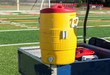 Yellow water cooler on a blue cart with soccer net in background