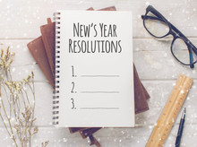 Notebook With New's Year Resolutions Massage, Glasses And Working Ornament On White Wooden Table Background.