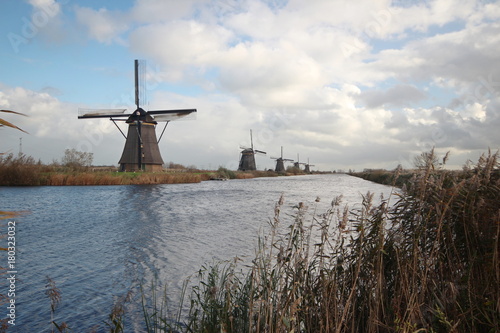 Plakat Dziejowy wiatraczek przy Kinderdijk terenem w holandiach z niebieskim niebem i dużymi białymi chmurami