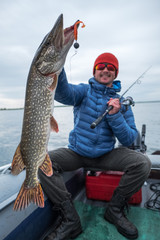 Wall Mural - Happy angler holds northern pike (Esox lucius), fish is around 2.3 kg (5lb). Fisherman sits in a boat with river on the background