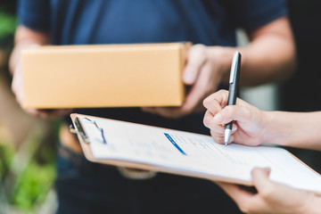 Close up shot of woman hand putting signature in clipboard to receive package from delivery man in background