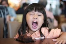 Asian Children Cute Or Kid Girl Enjoy And Fun With Happy Eating Delicious Brownie Chocolate Cake For Sweet Dessert Or Snack On Wood Table And Open Mouth At Lunch In Restaurant Or Cafe For Background