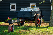 Vintage Cart With Dairy Milk Cans In Front Of Traditional Old Wooden House