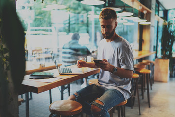 Young handsome hipster guy searching information online on a modern laptop and  checking email box, chatting in social network on mobile phone while sitting in cafe and using 5G internet connection.