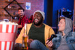 © Yakobchuk Olena - Playing games together. Stylish relaxed playful international friends are sitting on couch while enjoying home video console and having fun. Bearded guy is resting in background. Popcorn in foreground