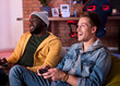 Excited players. Cheerful stylish young man is sitting on couch and using home video console. He is expressing gladness while looking at screen. Positive african guy is holding joystick in background