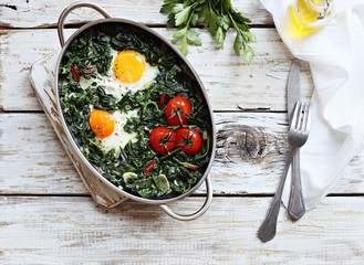 Eggs baked with spinach and tomatoes in serving pan overhead rustic wooden table. Overhead view.