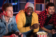 © Yakobchuk Olena - Rest with friends. Portrait of positive trendy african guy is sitting on couch and eating popcorn while looking at camera with smile. Two young concentrated men are playing video game. Selective focus