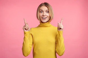 People and advertisment concept. Happy attractive woman wears yellow sweater, points up with fore fingers as shows something, has surprised pleased expression. Female gestures over pink wall