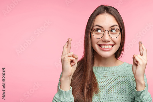 Fotografía Beautiful female student prays before exam, crosses fingers, hopes for good luck, has great desire to recieve excellent mark, smiles pleasantly, isolated over pink background with copy space