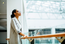 Young African Business Woman In Glasses Dressed In Gray Suit Holding On Handrail When Looking Up And Thinking Indoor At Modern Business Center. People Lifestyle Concept.