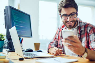 Young web developer working at his desk and checking his phone