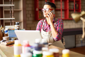 Wall Mural - Portrait of creative young woman working in art studio managing clients, using laptop and speaking by phone sitting at wooden table