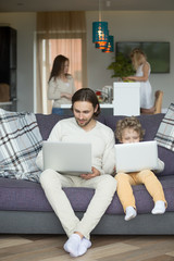 Wall Mural - Dad and boy using laptops on sofa, kid imitating copying daddy holding computer sitting on couch while mother with daughter cooking spending time together at home, like father like son, vertical