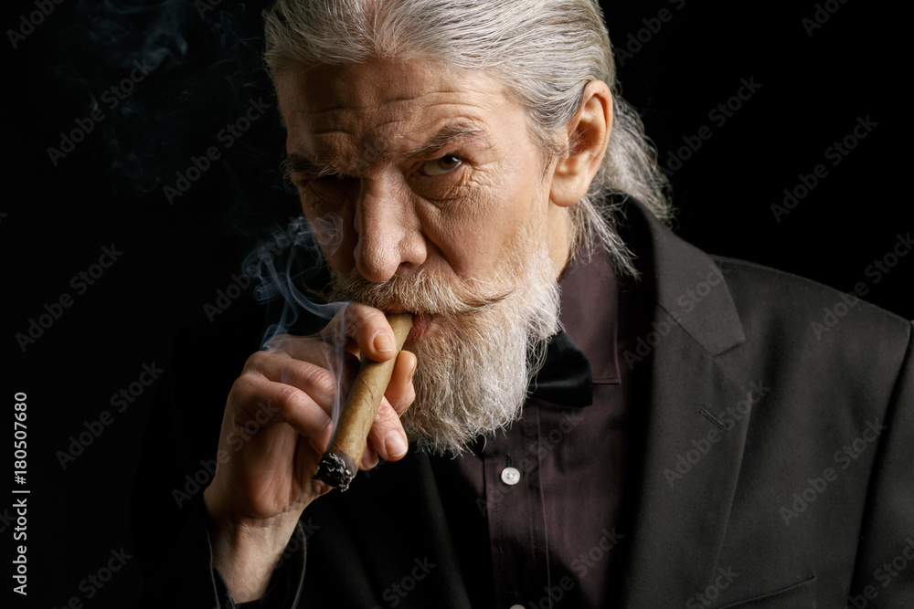 Stylish old man smoking cigar. Aged male in black suit in studio. Stock  Photo