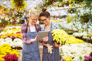 Portrait of two happy modern florist girls with eyeglasses and tablet in the greenhouse full of flowers watching video tutorials on the internet.