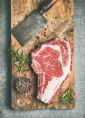 Wall Mural - Flat-lay of raw prime beef meat dry-aged steak rib-eye on bone and chopping knife on wooden board over grey concrete background with seasoning, top view. Meat high-protein dinner concept