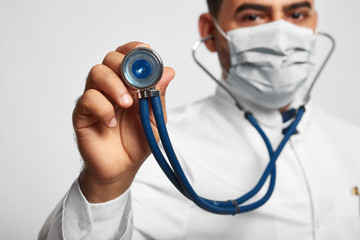 Selective focus on a stethoscope in the hand of a male doctor posing confidently at studio wearing protective mask people healthcare medicine scrubs occupation living equipment examination concept.