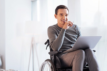 Having secret. Enigmatical male keeping smile on his face and holding glasses in right hand while sitting on the wheelchair