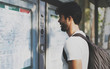 Young bearded interantional student searching the route on a public transportation system map in unknown city. Handsome traveler with backpack looking at train timetable on a railway station.