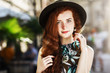 Outdoor close up portrait of young beautiful redhead girl with freckles, long curly hair. Model posing in street, wearing stylish hat, summer dress. Female beauty, fashion concept. Copy, empty space