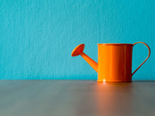 Orange Watering Cans Put On Wooden Table. The Background Is Turquoise And Copy Space For Content.
