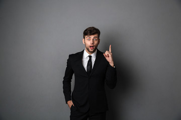 Poster - Amazed young man in suit pointing with finger upward, looking at camera