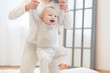© Rithor - Cute little baby is smiling while learning to walk, mom is holding his hands. first steps of child. White light domestic interior, white clothes