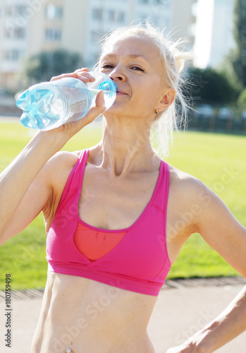 Poster Blond slim sportswoman is drinking water from the bottle on the stadium