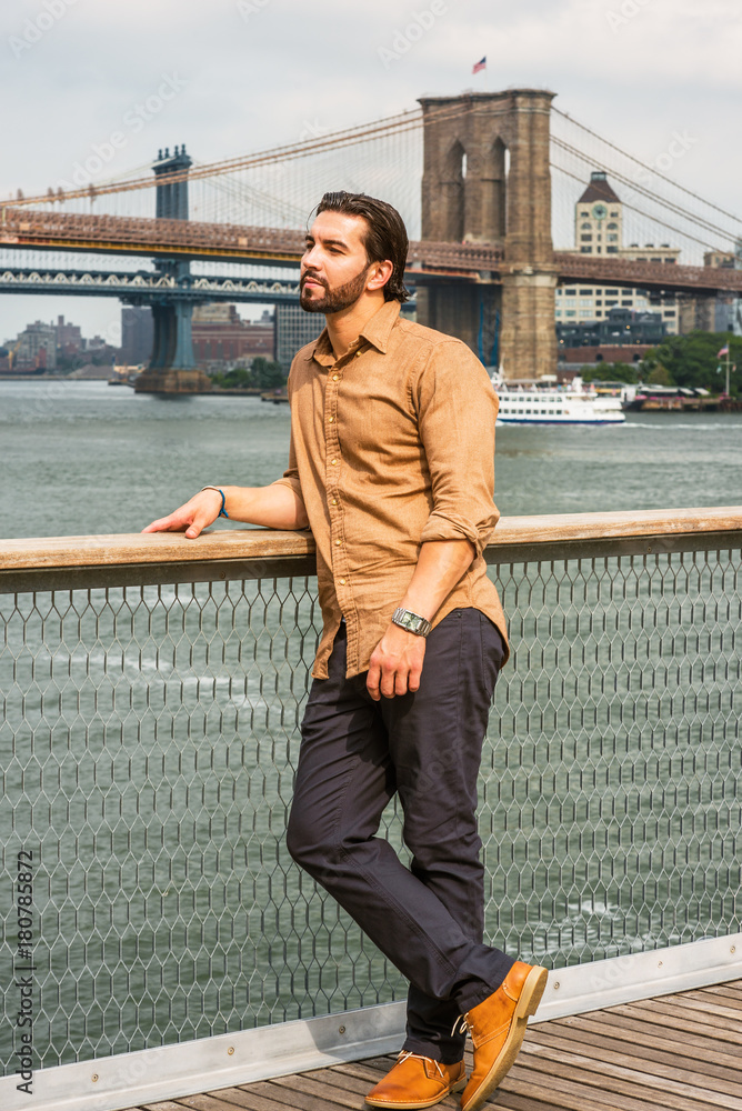 Photo Stock American Man with beard traveling in New York, wearing brown  shirt, black pants, brown leather shoes, standing by fence at harbor,  thinking. Manhattan, Brooklyn bridges, boats on background.. | Adobe