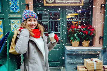 Attractive cute girl in with the shopping bags and holding disposable cup with warm drink in her hand. Female dressed in warm bright hat, red scarf and mittens. Happy holiday and freedom concept.