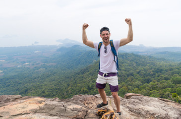 Wall Mural - Tourist Man With Backpack Standing On Mountain Top Raised Hands Happy Smiling Over Beautiful Landscape, Young Guy On Hike