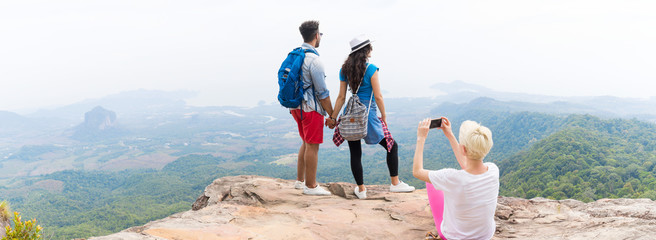 Wall Mural - Girl Taking Photo Of Couple With Backpacks Posing Over Mountain Landscape On Cell Smart Phone Panorama, Trekking Young Man And Woman Group On Hike Tourists Adventure Activity