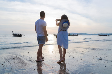Wall Mural - couple beach summer vacation, man woman take photo sunset young guy girl back rear view sea ocean ho