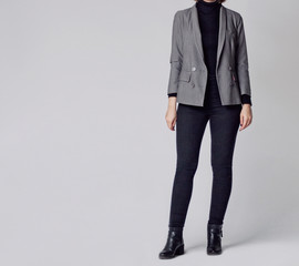 Poster - Woman wearing stylish and smart outfit with black turtleneck, gray elegant double-breasted blazer, black skinny jeans and black ankle boots isolated on gray background. Copy space